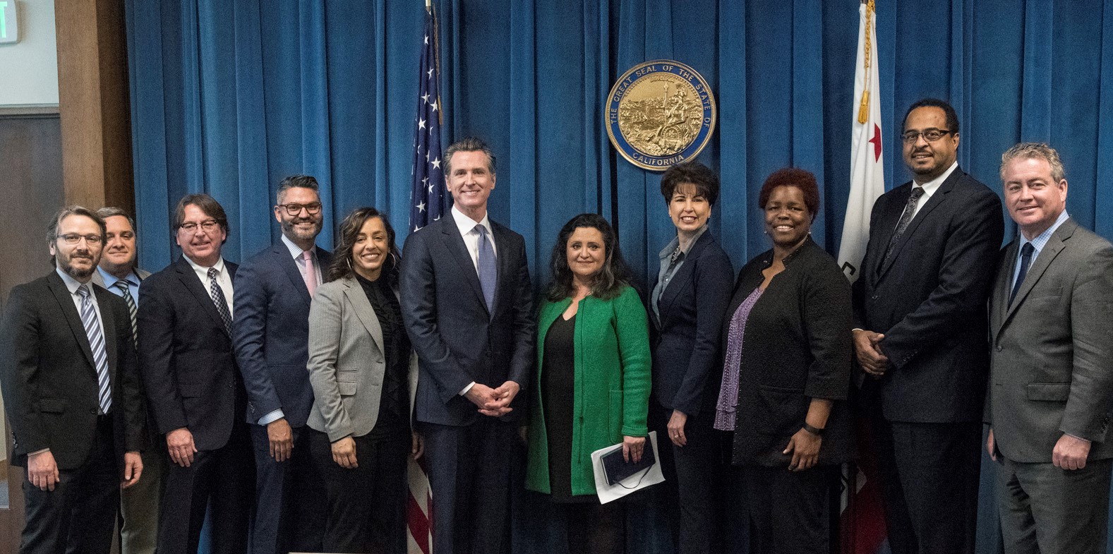 Governor Gavin Newsom standing with ten other officials in business attire, posing for a group photo in front of the California State Seal and American and California flags.