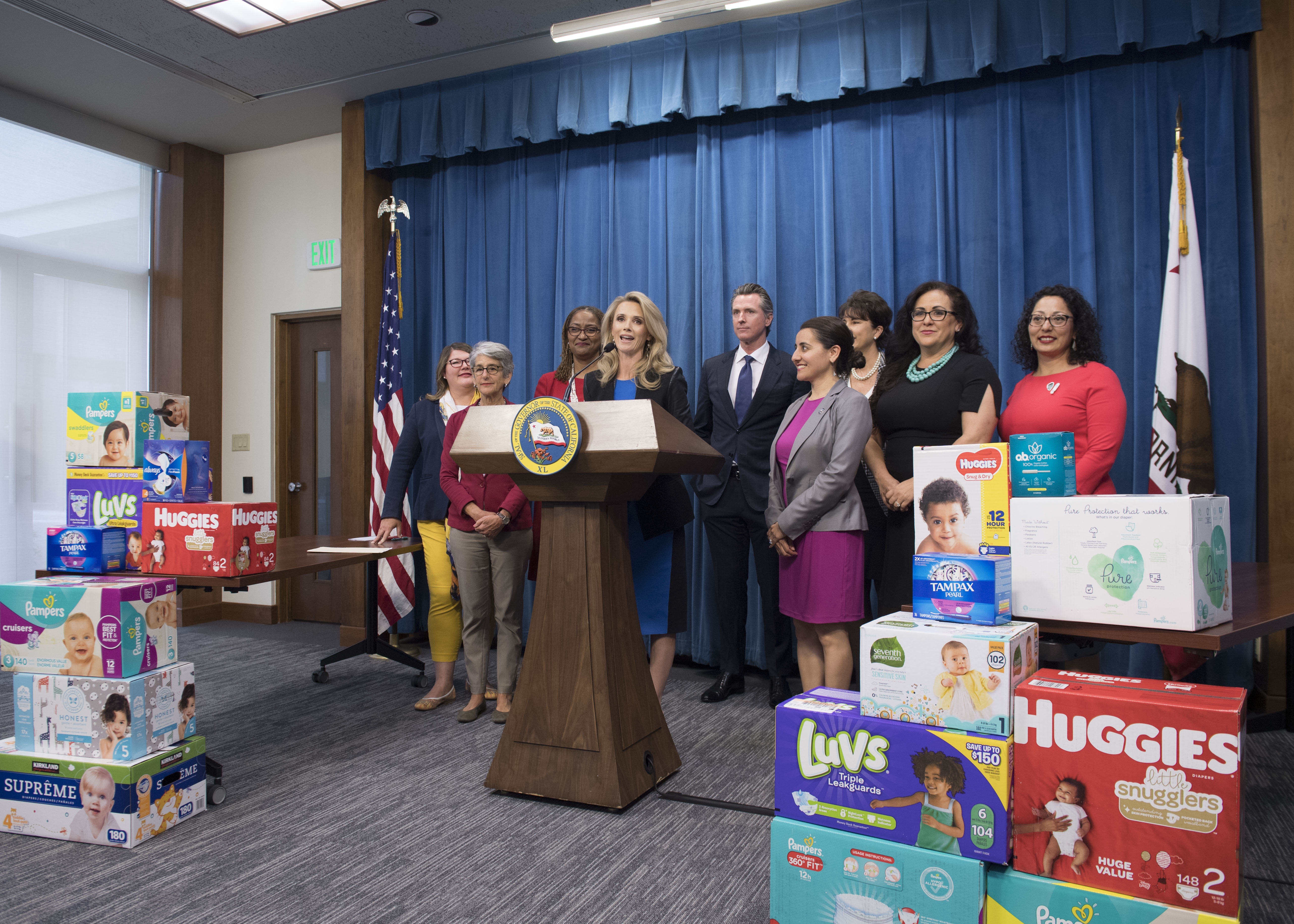 Press conference at a podium bearing the seal of the Governor of California, with First Partner Jennifer Siebel Newsom speaking at the microphone flanked by a group of officials including Governor Newsom, surrounded by stacked boxes of diapers and feminine hygiene products from brands like Pampers, Huggies, Luvs, and Tampax, with American and California flags in the background.