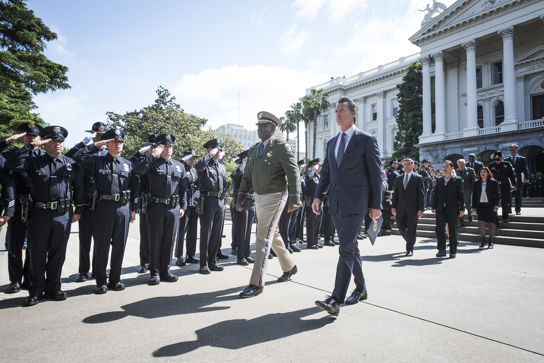 Governor Newsom walks alongside a uniformed CHP official past a formation of saluting law enforcement officers in front of the California State Capitol building, with other officials following behind on the plaza.