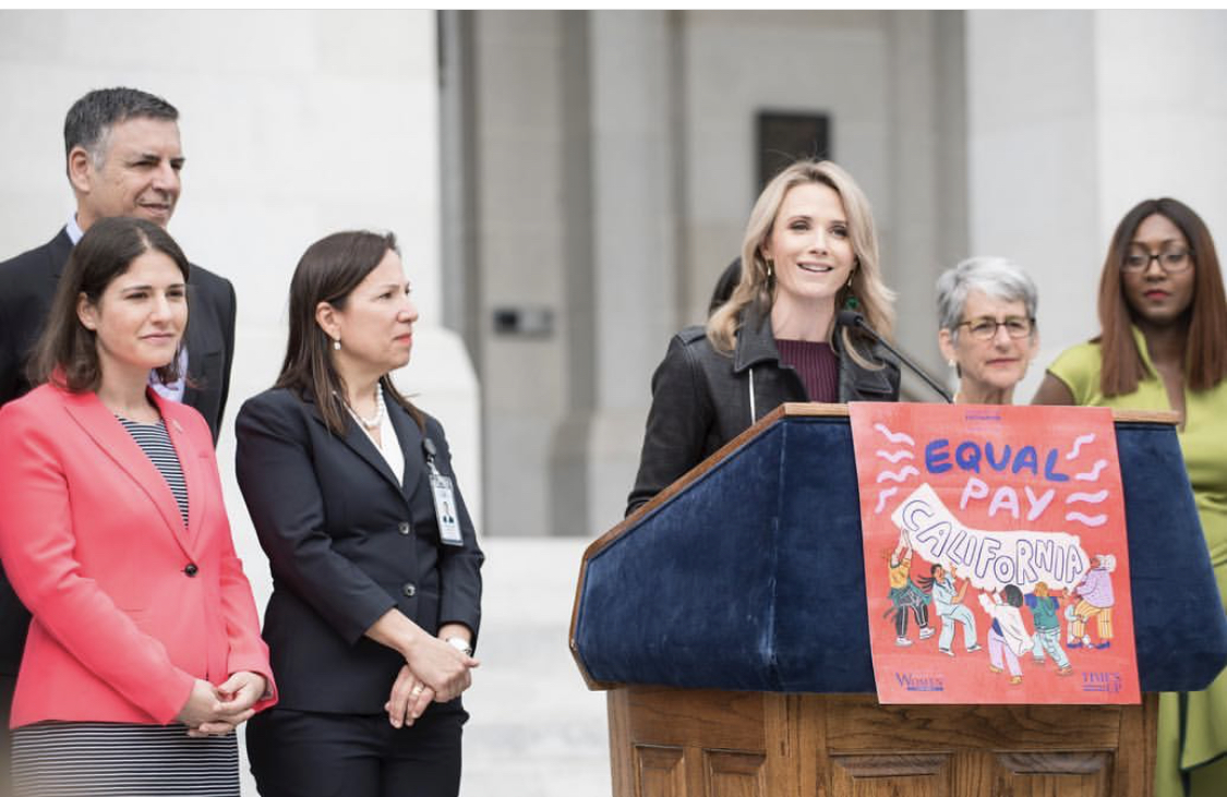 Jennifer Siebel Newsom speaks at a wooden podium decorated with an "Equal Pay California" illustrated poster, flanked by a group of officials on the steps of a stone government building.