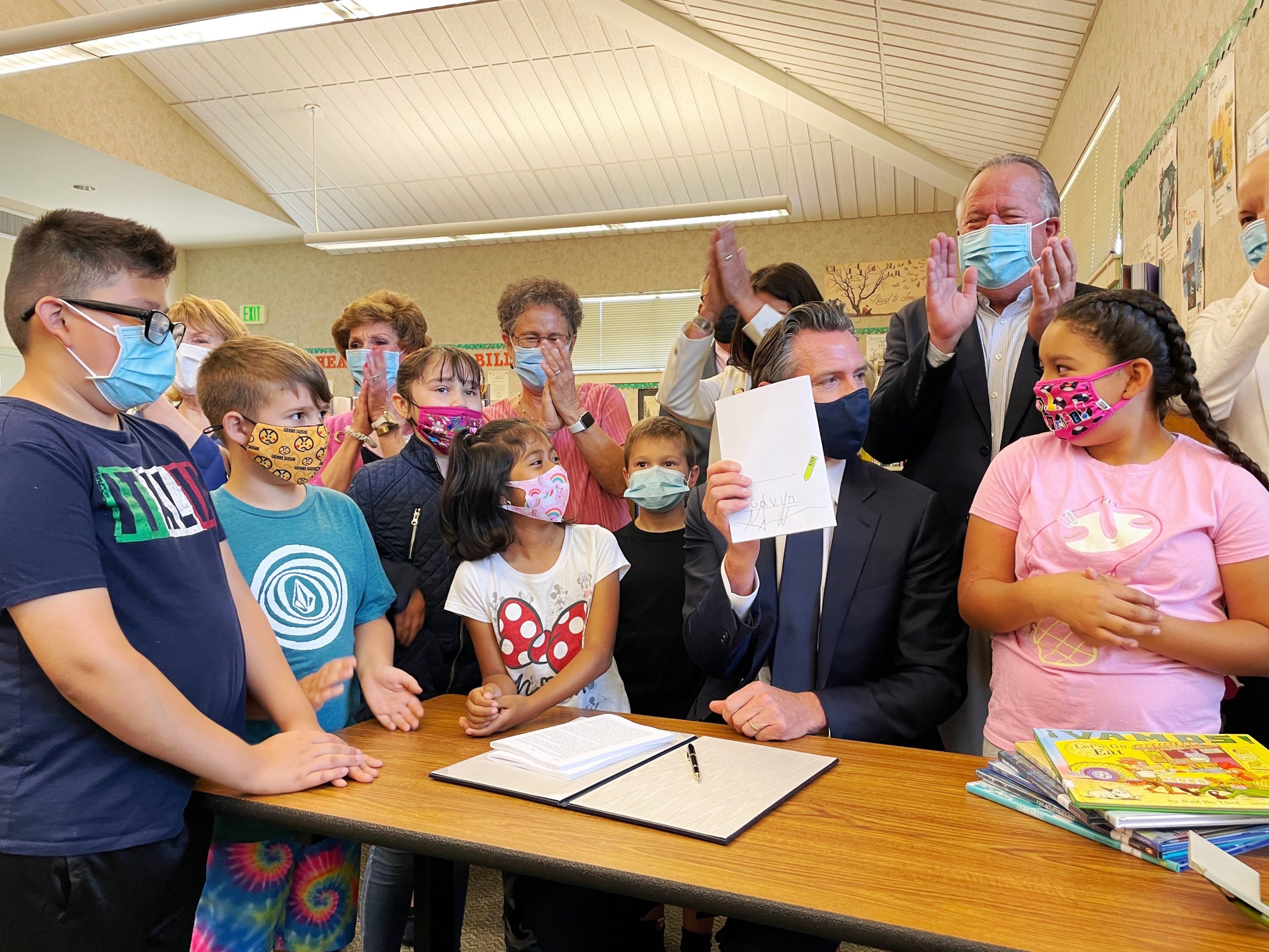 Governor Gavin Newsom sits at a table in a school setting holding up the signed state budget law.  He is surrounded by children and adults clapping.