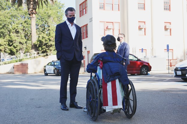 Governor Gavin Newsom, wearing a face mask, speaking with a person in a wheelchair on a street, with another masked man and parked cars in the background.