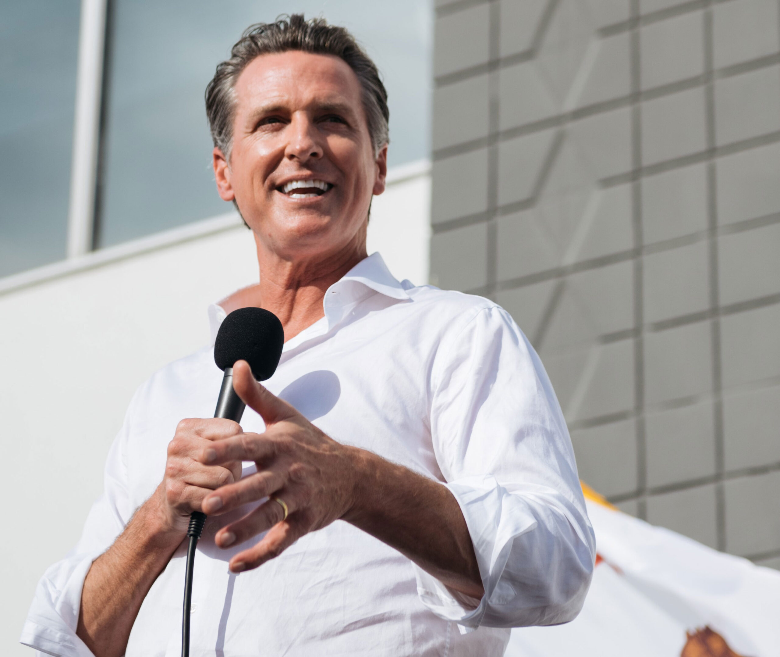 Governor Gavin Newsom, a medium skin toned white man with salt and pepper hair, wears a white button down shirt with rolled sleeves smiles while holding a microphone. A part of the California flag is in the background.