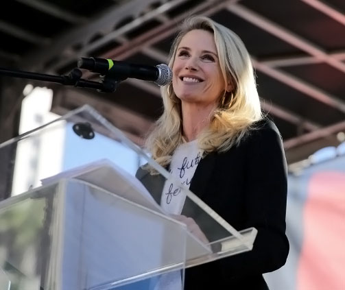 First Partner Jennifer Siebel Newsom, a light skin toned woman with blonde hair smiles from behind a podium outside.