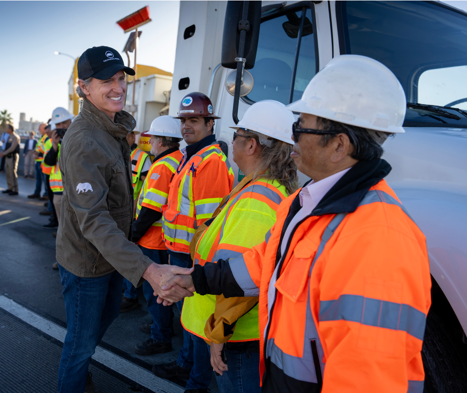 Governor Newsom wears an olive green utility jacket and smiles as he shakes the hand of a CalTrans worker. Workers in hardhats and reflective gear line up in front of a large work truck to shake the Governor’s hand.