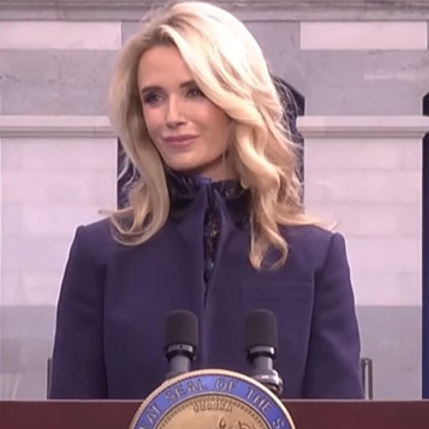 Wearing a formal navy blazer, First Partner Jennifer Siebel Newsom smiles from behind a podium at the California Republic Inauguration. On the front of the podium in the state seal.