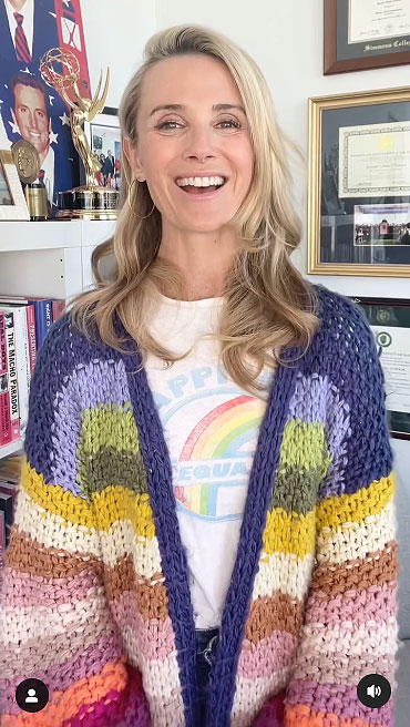 First Partner Jennifer Siebel Newsome smiles widely while wearing a graphic t-shirt with a faded rainbow and a colorful striped knitted cardigan.