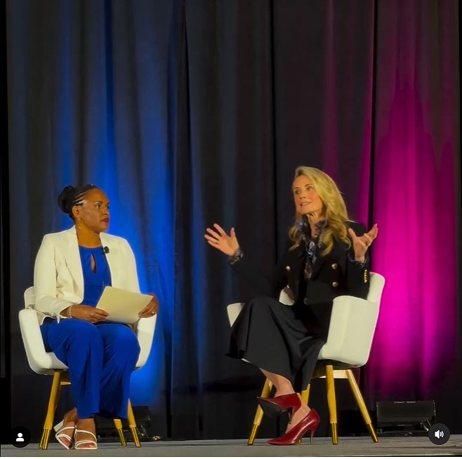 First Partner Jennifer Siebel Newsom and Kimberly Ellis sit on a stage during a talk. Behind them are stage curtains with blue and dark pink lighting on them.