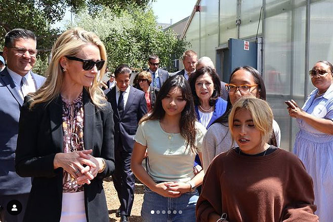 First Partner Jennifer Siebel Newsom smiles and listens as she walks with a group of female students outside. A group of adult professionals walk behind them.