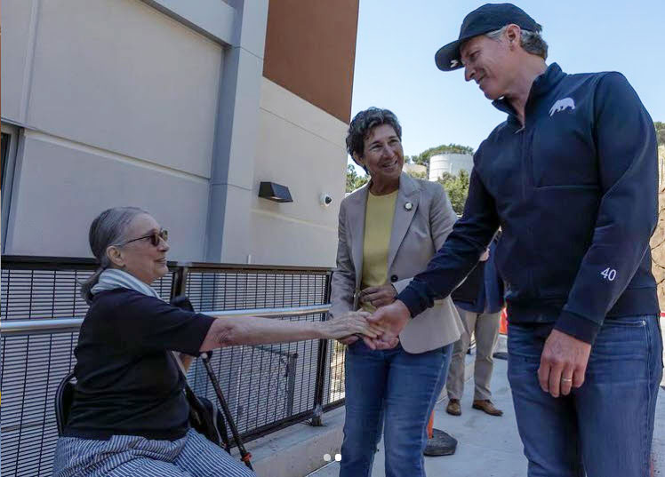 Outside of a building, Governor Gavin Newsom shakes the hand of an older woman that is seated. A woman smiles as they shake hands.