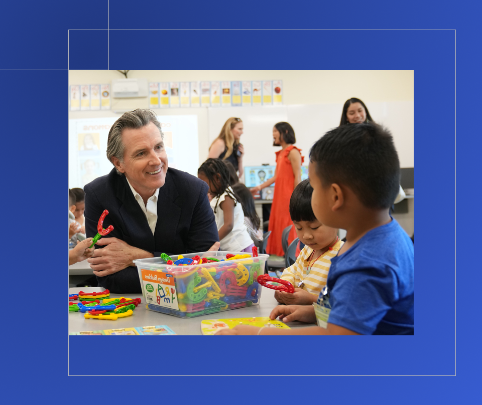Governor Newsom smiles at a young child at they sit at a table in a classroom and play with colorful learning materials.