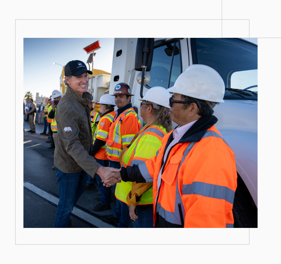 Governor Newsom wears an olive green utility jacket and smiles as he shakes the hand of a CalTrans worker. Workers in hardhats and reflective gear line up in front of a large work truck to shake the Governor’s hand.