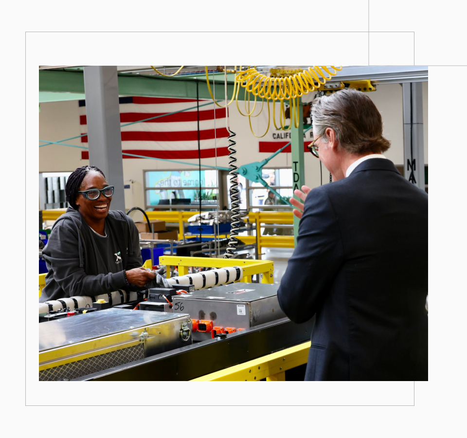 Governor Newsom tours a power factory and shares a smile with a brown skin-toned woman in protective glasses.