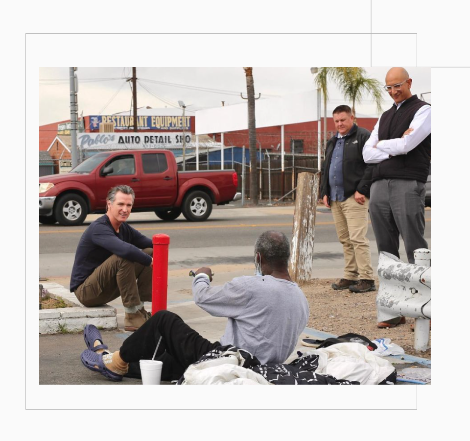 Governor Gavin Newsom sits on a curb in Fresno as he talk to an older homeless man with a dark skin tone who is sitting on a sleeping bag. Two men observe their interaction.