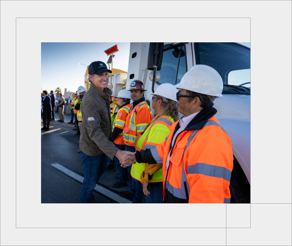 Governor Newsom wears an olive green utility jacket and smiles as he shakes the hand of a CalTrans worker. Workers in hardhats and reflective gear line up in front of a large work truck to shake the Governor’s hand.