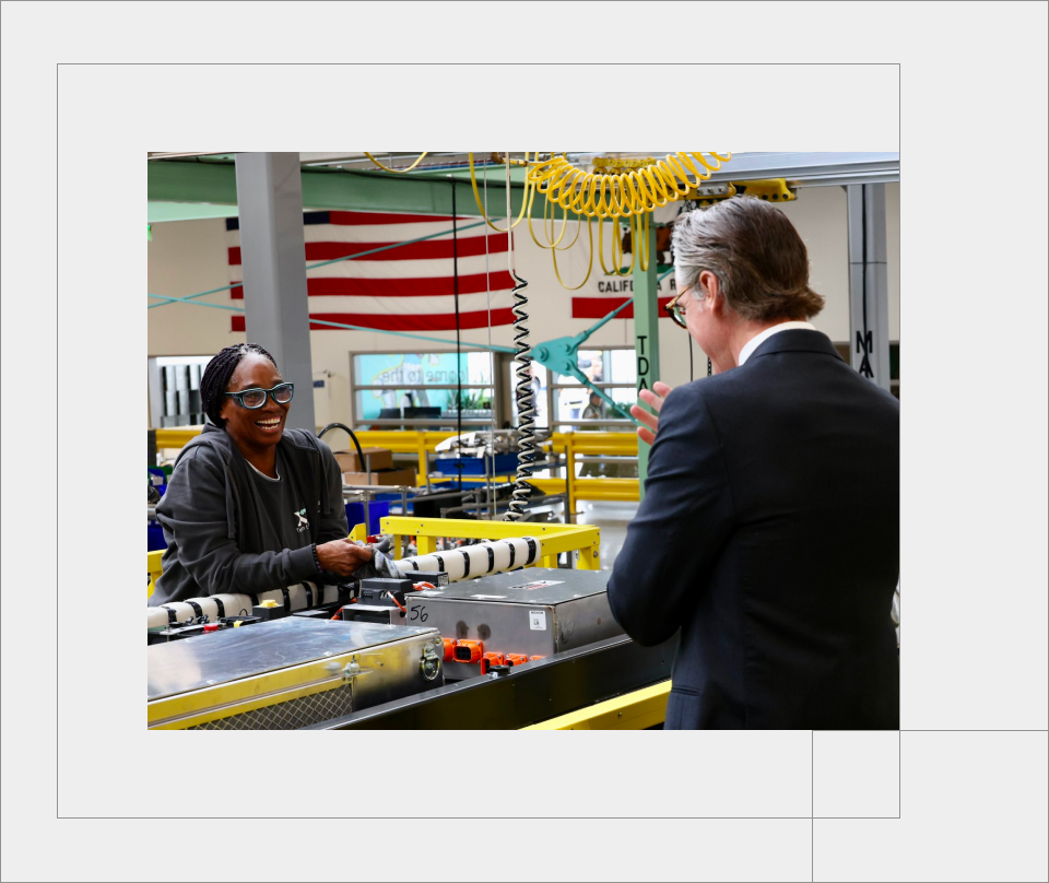 Governor Newsom tours a power factory and shares a smile with a brown skin-toned woman in protective glasses.