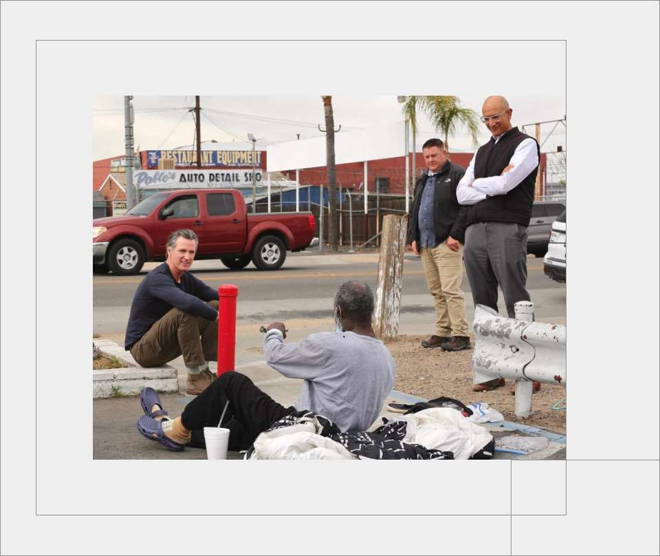 Governor Gavin Newsom sits on a curb in Fresno as he talk to an older homeless man with a dark skin tone who is sitting on a sleeping bag. Two men observe their interaction.