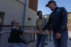Governor Gavin Newsom, wearing a cap and California bear-logo jacket, leans down to shake hands with a seated older woman as a woman in a beige blazer smiles beside him outside a building.