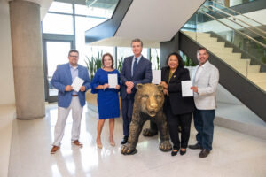 Photo of Governor Newsom standing with four other people around a bronze bear statue in a lobby, several holding signed documents, with a staircase and large windows visible in the background.