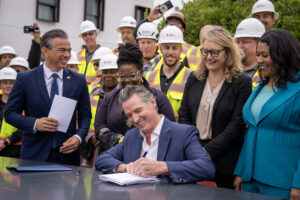 Photo of Governor Newsom seated at a table, smiling while signing a document with a pen, surrounded by a group of smiling officials and construction workers wearing hard hats and safety vests, outdoors in front of a building.