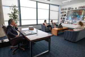Governor Newsom sits at his desk in his office listening to a video call displayed on a wall-mounted screen, while three staff members sit on couches nearby.