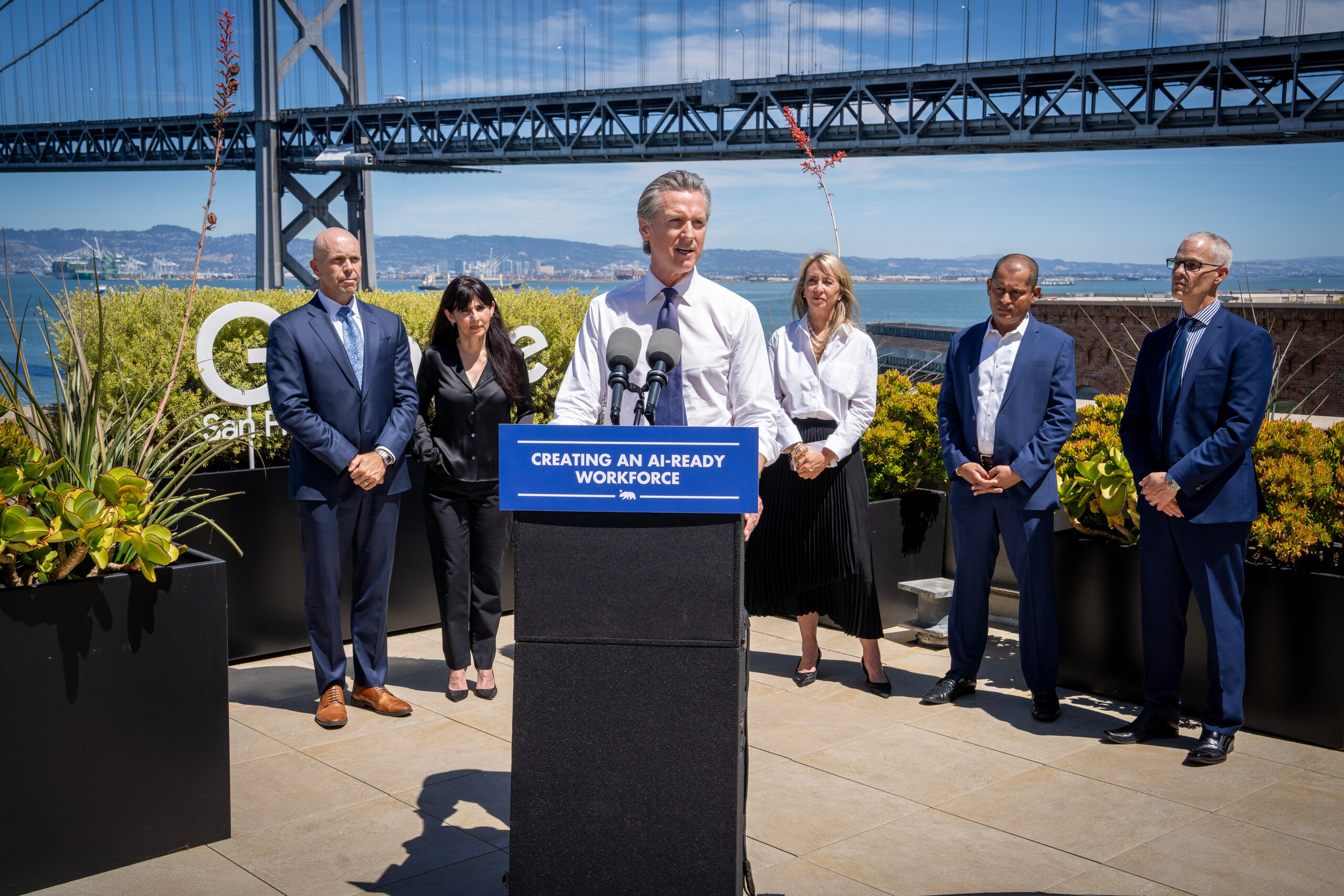 A man in a white dress shirt and tie speaks at a podium with a blue sign reading “Creating an AI-Ready Workforce,” while five people in business attire stand behind him on a rooftop terrace with the Bay Bridge and water in the background.
