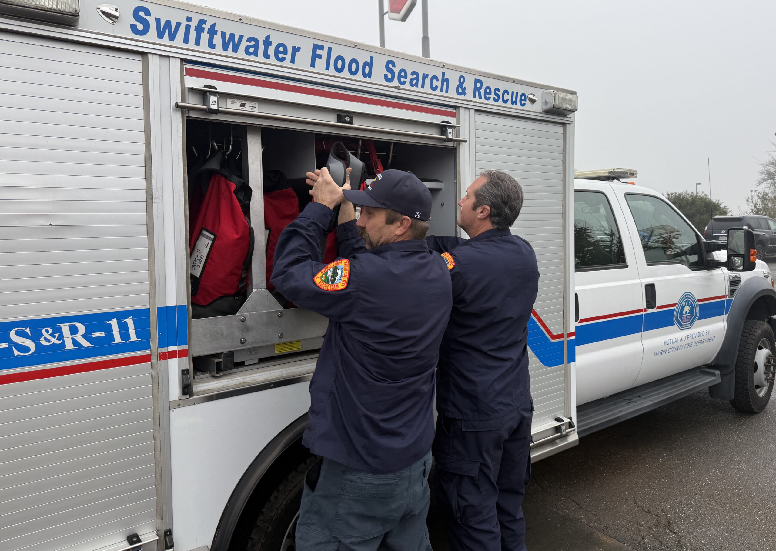 Two uniformed emergency responders stand beside a “Swiftwater Flood Search & Rescue” truck, retrieving red rescue gear from an open storage compartment.