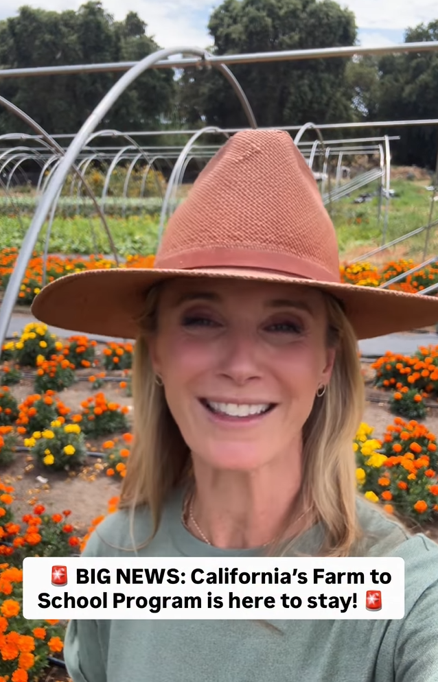 Selfie-style photo of First Partner Jennifer Siebel Newsom wearing a wide-brimmed hat, smiling in a field of orange and yellow marigolds with greenhouse structures in the background, with on-screen text reading Big News, California's Farm to School Program is here to stay.