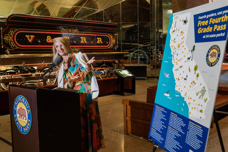 First Partner Jennifer Siebel Newsom in a colorful patterned outfit speaks at a podium bearing the California State Parks logo, standing in front of a vintage locomotive labeled 'V. & T.R.R.' inside a museum. A sign to the right advertises the 'Free Fourth Grade Pass' program with a map of California state parks.