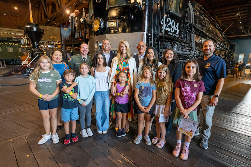 Group photo of adults including First Partner Jennifer Siebel Newsom and children posing together on a wooden platform in front of a large black locomotive numbered 4294, inside a train museum.