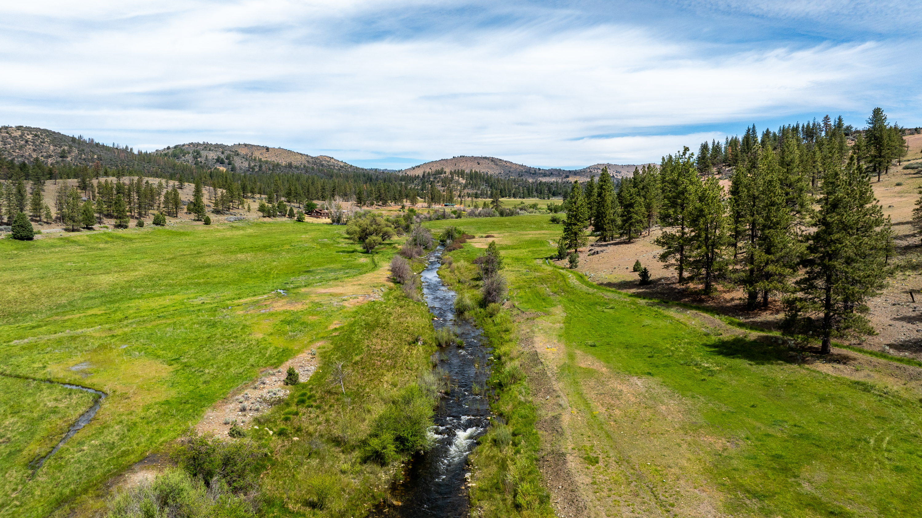 Aerial photo of a winding creek running through a green valley meadow, flanked by pine trees and low sagebrush-covered hills. A small cabin sits near the tree line in the distance, with rolling mountains under a partly cloudy sky in the background.