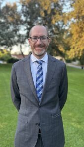 Professional photo of a smiling man with glasses and a shaved head, wearing a gray plaid blazer, light blue shirt, and striped tie, standing outdoors on a lawn with trees and a park path in the background.