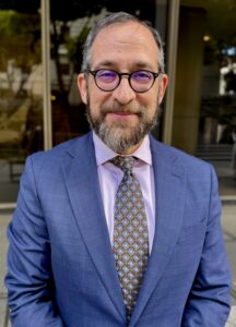 Professional photo of a smiling man with gray curly hair, a beard, and round glasses, wearing a blue suit, light purple shirt, and patterned tie, standing outdoors in front of a glass building.