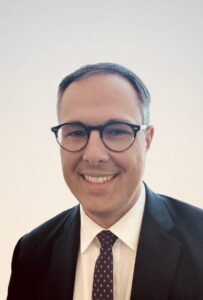 Professional headshot of a smiling man with dark hair and glasses, wearing a black suit, white shirt, and a dotted tie, photographed against a plain light beige background.