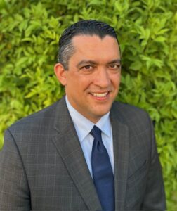 Professional headshot of a smiling man with dark hair, wearing a gray plaid suit with a light blue shirt and dark blue tie, photographed against a green hedge background.