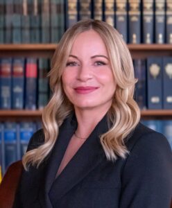 Professional headshot of a smiling woman with long wavy blonde hair, wearing a black blazer, photographed in front of a bookshelf filled with matching navy law volumes.