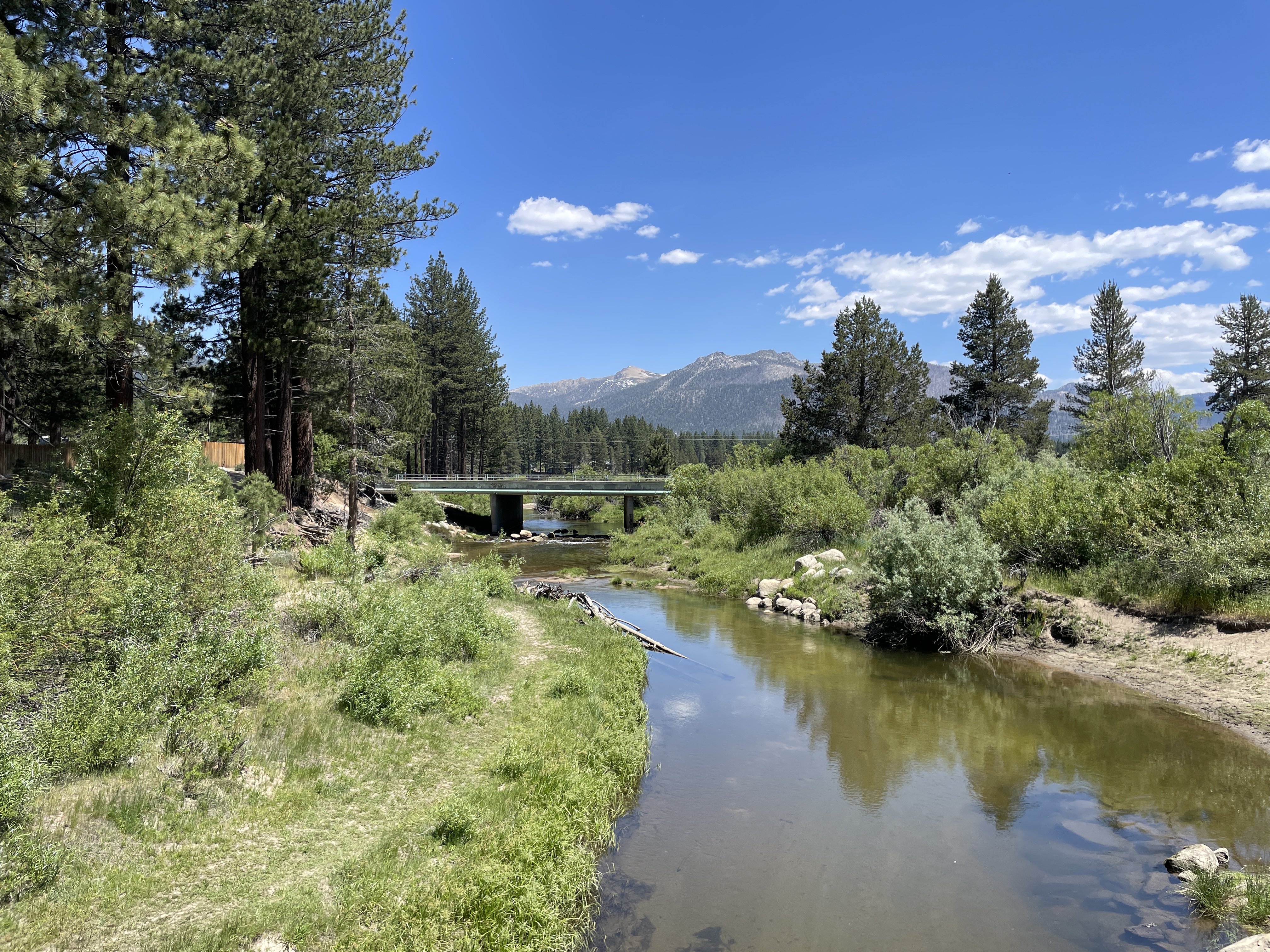 Ground-level photo of a calm river reflecting the blue sky, flowing under a small footbridge and lined with pine trees and green shrubs. Jagged mountain peaks rise in the distance under a partly cloudy sky.