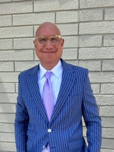 Photo of a smiling bald man wearing glasses, a navy pinstriped suit, white shirt, and light purple tie, standing in front of a light gray cinder block wall.