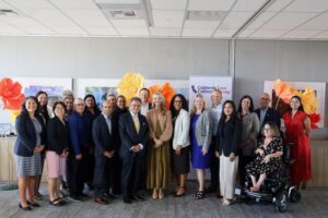 Group photo of about twenty people with First Partner Jennifer Siebel Newsom in business attire standing together in an indoor event space, smiling for the camera. Large decorative paper poppy flowers in orange and yellow frame the group, and a banner reading 'California Love, California Strong' is visible in the background alongside framed photos on the wall.