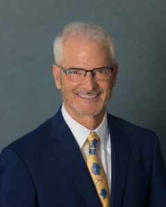 Professional headshot of a smiling man with white hair and glasses, wearing a navy suit, white shirt, and a yellow patterned tie, photographed against a muted teal-gray background.
