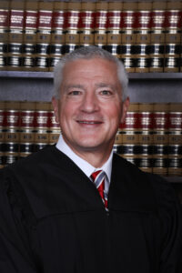 Professional portrait of a smiling man with gray hair wearing a judge's black robe over a white shirt and red striped tie, photographed in front of a bookshelf filled with matching law volumes.