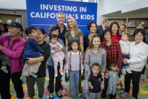 Group photo of Governor Newsom, First Partner Jennifer Siebel Newsom, adults, children, and toddlers posing together for a large photo in front of a blue 'INVESTING IN CALIFORNIA'S KIDS' banner, with several people smiling broadly, standing on a colorful alphabet play mat in a room with bookshelves and children's toys.