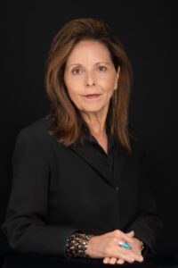 Professional headshot of a woman with shoulder-length brown hair, wearing a black blouse and a beaded bracelet, hands clasped in front of her, photographed against a black background.