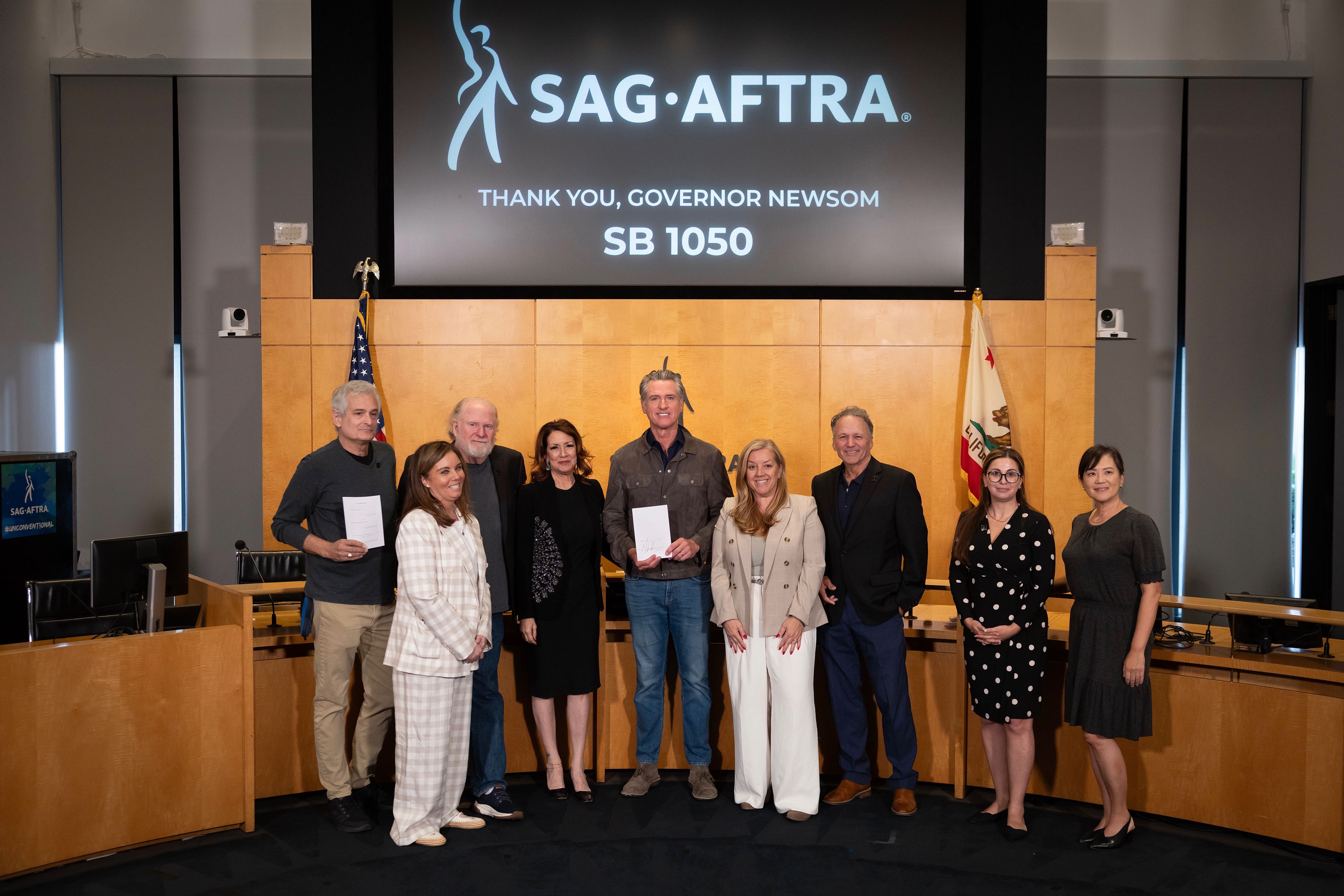 Governor Gavin Newsom holds signed legislation at SAG-AFTRA headquarters, joined by union leaders, and Senator Angelique Ashby (D-Sacramento), with the U.S. and California flags displayed behind them.
