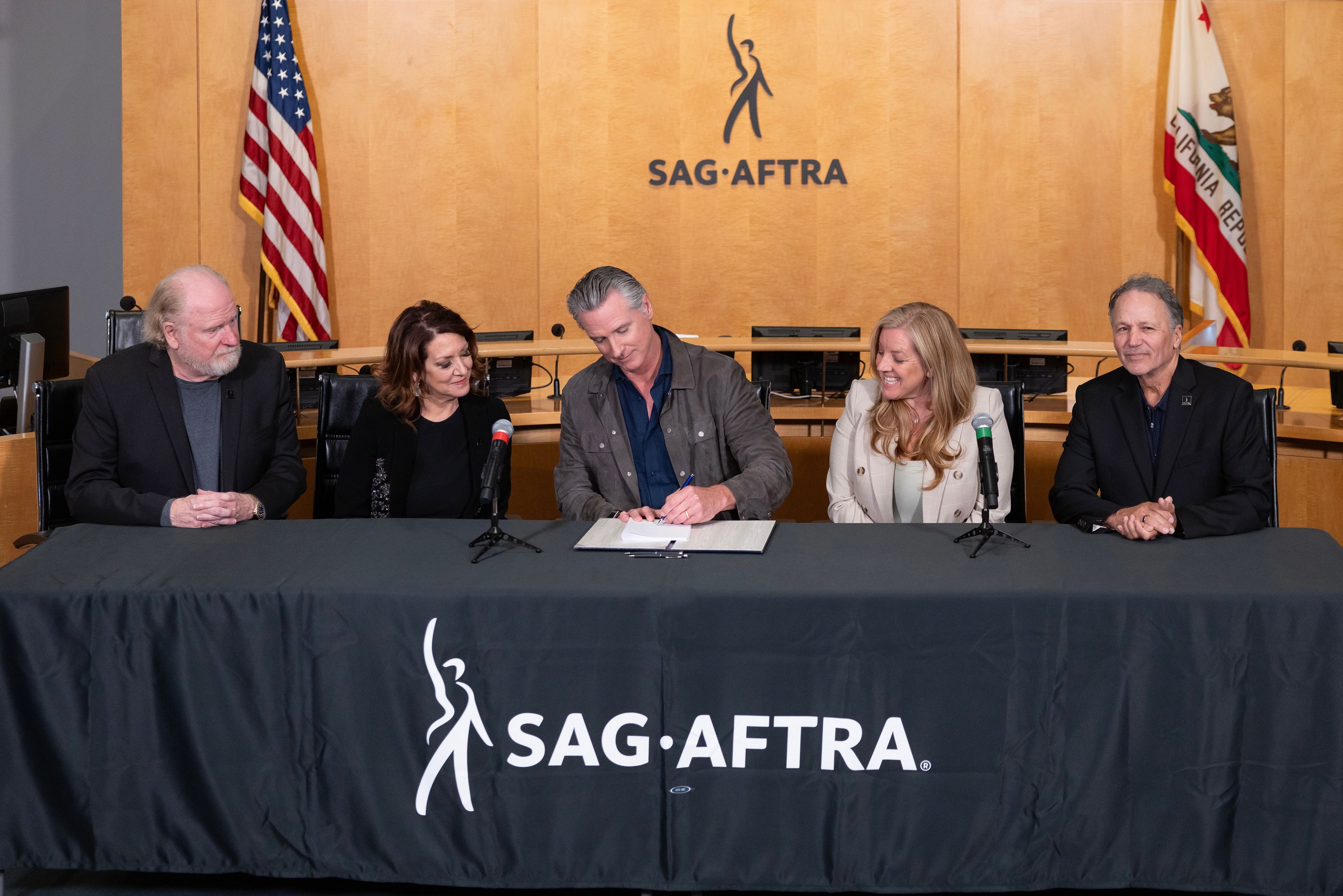 Governor Gavin Newsom signs legislation at SAG-AFTRA headquarters, joined by union leaders, and Senator Angelique Ashby (D-Sacramento), with the U.S. and California flags displayed behind them.