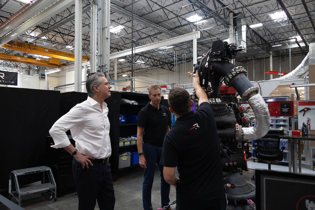 Three men stand around a large black rocket engine in a high-ceilinged manufacturing facility. One man in a white dress shirt looks on with a hand on his hip while a worker in a black polo reaches up to point at part of the engine, and another worker in a black polo watches nearby.