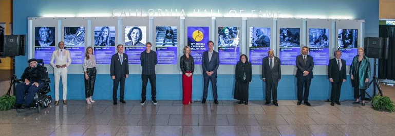 A diverse group of people stand side by side in front of the California Hall of Fame exhibit, with First Partner Jennifer Siebel Newsom and Governor Gavin Newsom standing in the middle. Behind the group are photos and plaques highlighting important figures in Californian history. Pictured are Guy Johnson representing Dr. Maya Angelou; RuPaul Charles; Brandi Chastain; Fred Córdova representing Dr. France A. Córdova; Tony Hawk; First Partner Siebel Newsom; Gov. Gavin Newsom; Jeanne Wakatsuki Houston; Rev. James M. Lawson Jr.; George Lopez; Wolfgang Puck; Helen M. Turley at the 2019 California Hall of Fame Induction Ceremony on Tuesday, December 10, 2019, in Sacramento, California.