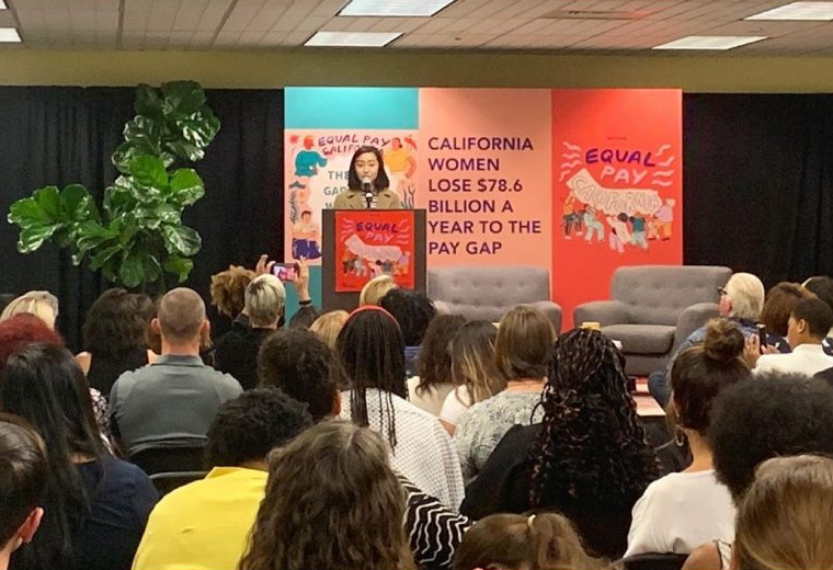 A woman is standing behind a podium in front of a crowd speaking about pay inequality in California. She is in front of a sign that says, "California women lose $78.6 billion a year to the pay gap."