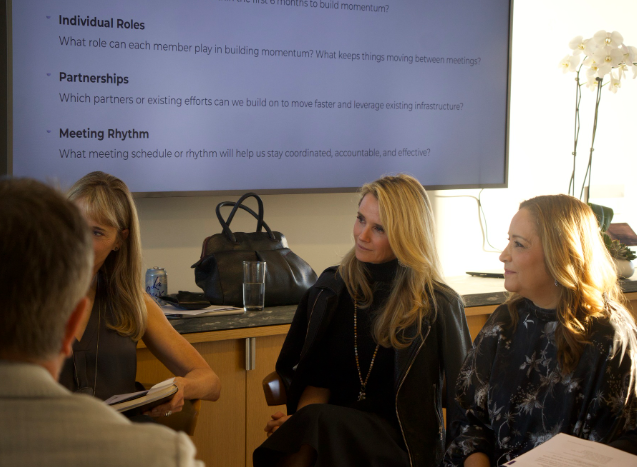 First Partner Jennifer Siebel Newsom pose with eight women by a bookshelf with framed awards and plaques. They are sitting around a round table with meeting notes.