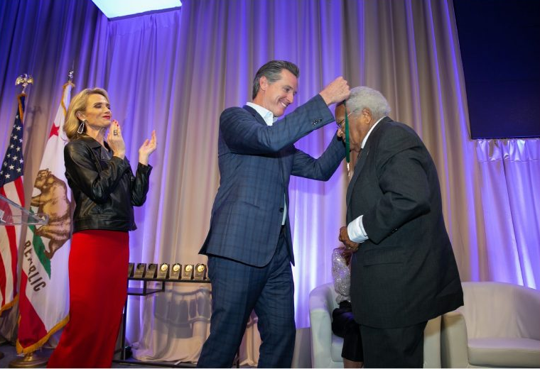 Governor Gavin Newsom, wearing a dark blue checkered suit, drapes a medal around the neck of an older man with darker skin, who is wearing a black suit. First Partner Jennifer Siebel Newsom is clapping in celebration behind Governor Newsom.